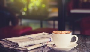 Weiße Kaffeetasse mit Untersetzer steht auf einem Tisch neben einer gefalteten Zeitung. Dahinter ist ein Fenster mit Blick auf den Garten.