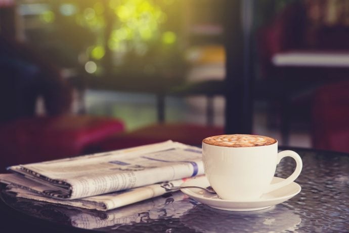 Weiße Kaffeetasse mit Untersetzer steht auf einem Tisch neben einer gefalteten Zeitung. Dahinter ist ein Fenster mit Blick auf den Garten.