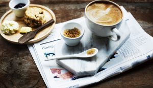 Auf einem Holztisch steht ein kleiner Frühstücksteller. Daneben steht auf einem Tablet eine Kaffeetasse mit Löffel und Zucker. Darunter liegt eine Zeitung.