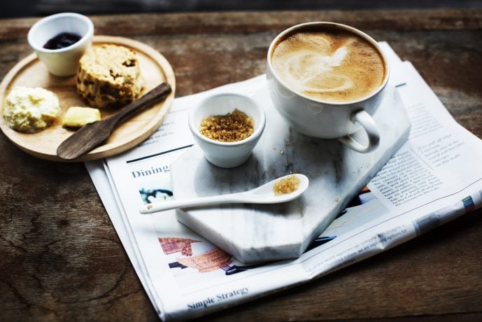 Auf einem Holztisch steht ein kleiner Frühstücksteller. Daneben steht auf einem Tablet eine Kaffeetasse mit Löffel und Zucker. Darunter liegt eine Zeitung.