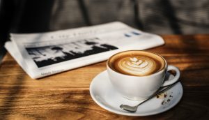 Eine weiße Kaffeetasse mit Untersetzer und Löffel steht auf einem Holztisch. Daneben liegt eine Zeitung.