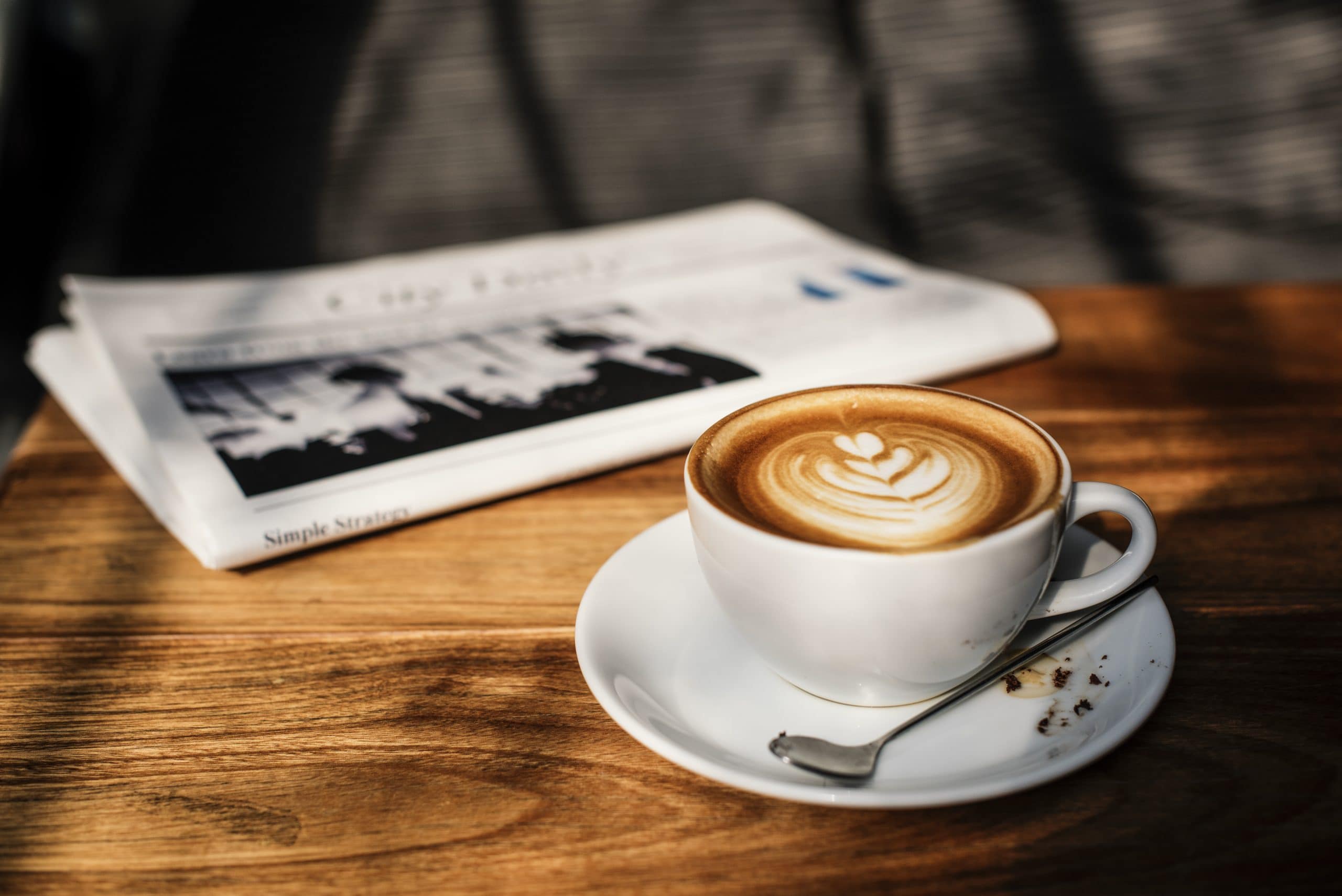 Eine weiße Kaffeetasse mit Untersetzer und Löffel steht auf einem Holztisch. Daneben liegt eine Zeitung.