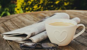 Eine weiße Kaffeetasse steht neben einer Zeitung und einer Brille auf einem Holztisch im Freien.