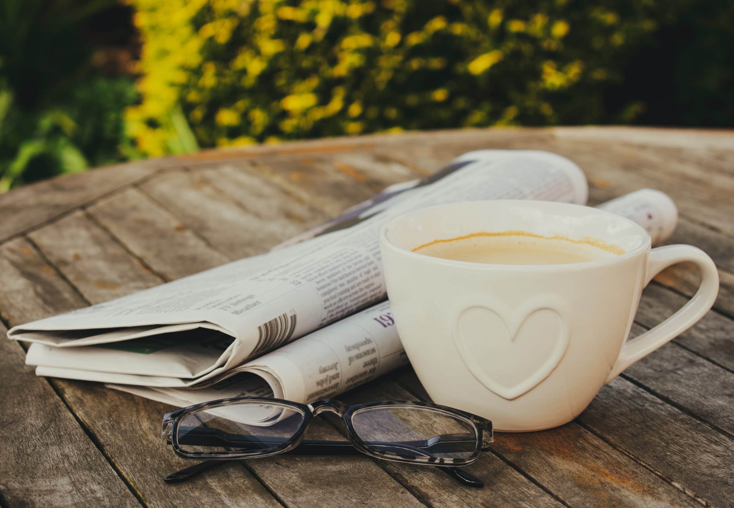 Eine weiße Kaffeetasse steht neben einer Zeitung und einer Brille auf einem Holztisch im Freien.