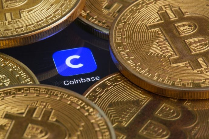 Ein Haufen Bitcoin liegt um ein Display, das das Logo der Coinbase-App zeigt.