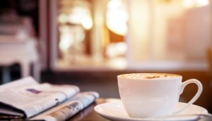 Kaffeetasse steht auf einem Untersetzer auf einem Tisch. Daneben liegen zwei Zeitungen übereinander.