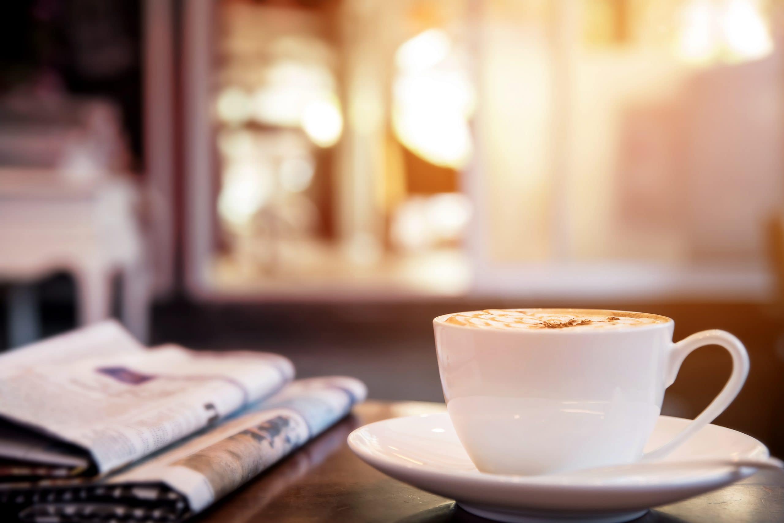 Kaffeetasse steht auf einem Untersetzer auf einem Tisch. Daneben liegen zwei Zeitungen übereinander.