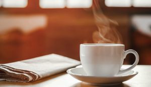 Eine Zeitung und eine Kaffeetasse samt Untersetzer stehen auf einem Tisch. Dahinter fällt Licht durch ein Fenster in den Raum.