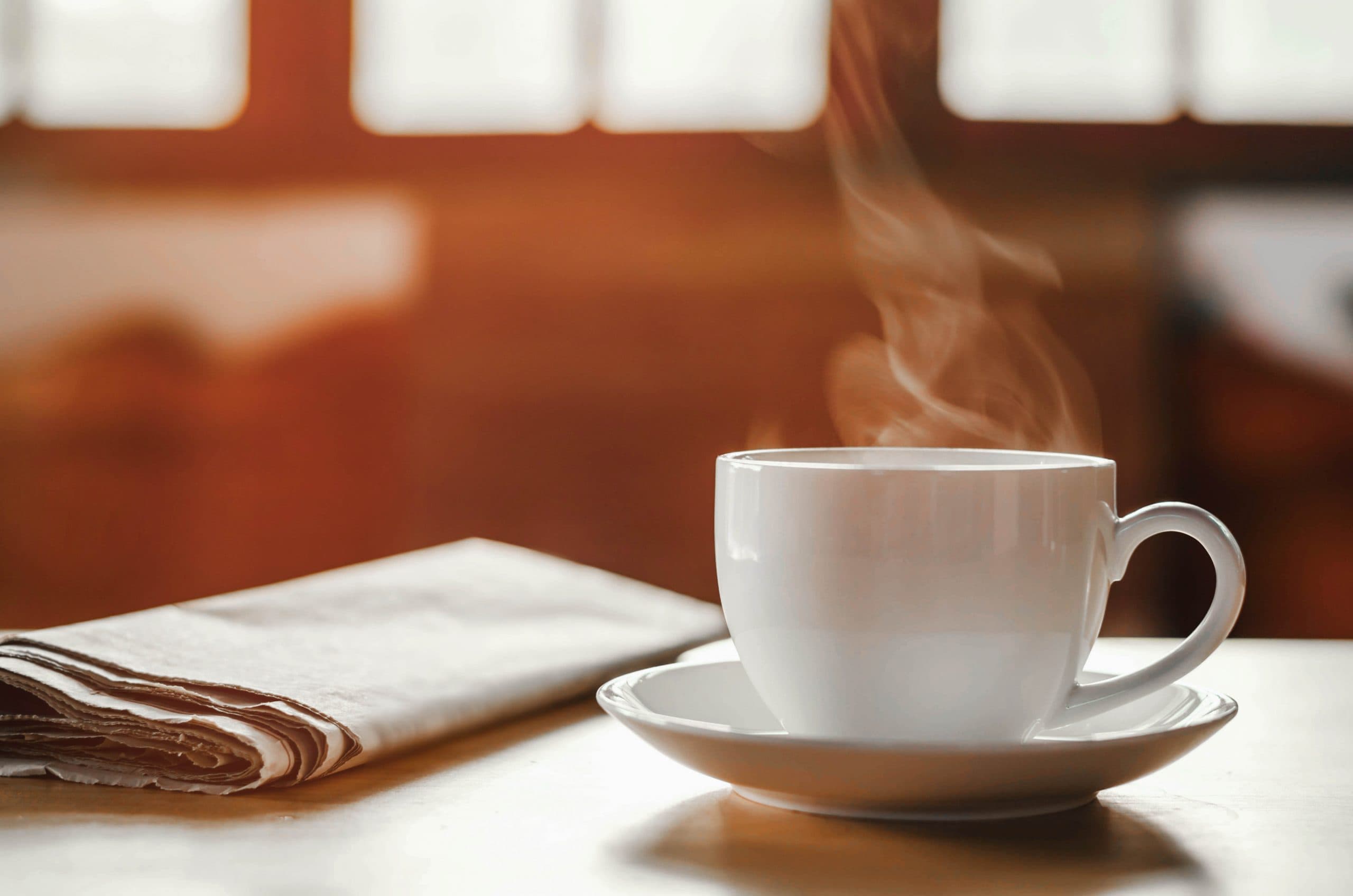 Eine Zeitung und eine Kaffeetasse samt Untersetzer stehen auf einem Tisch. Dahinter fällt Licht durch ein Fenster in den Raum.