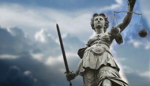 Justizia steht im Vordergrund, dahinter ist ein Wolkenreicher Himmel zu sehen.