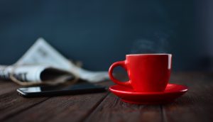Eine Zeitung, ein Handy und eine rote Tasse stehen auf einem Holztisch.