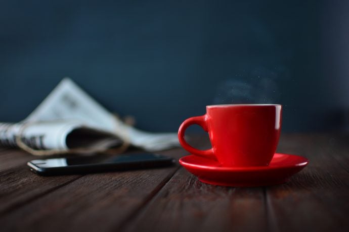 Eine Zeitung, ein Handy und eine rote Tasse stehen auf einem Holztisch.