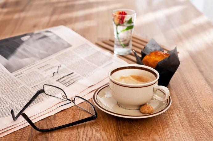 Eine Zeitung, eine Brille, eine Kaffeetasse samt Untertasse, ein Muffin und eine Blume stehen auf einem Holztisch.