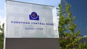 Eine gläserne Tafel mit dem Logo der EZB drauf. Daneben ist der Banken-Tower der Zentralbank.