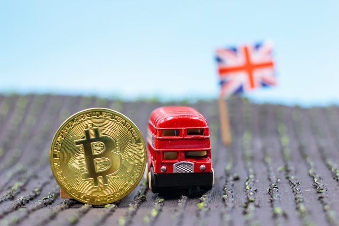 Bitcoin-Münze neben einem Londoner Spiezleugbus, im Hintergrund die Großbritannien-Flagge