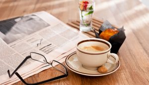 Eine Brille, eine Zeitung, eine Kaffeetasse, ein Muffin und eine Blume stehen auf einem Holztisch.