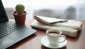 Zeitung, Kaktus, Laptop und Kaffeetasse stehen auf eine Holztisch.