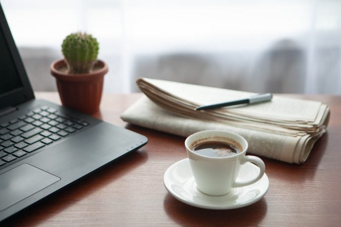 Zeitung, Kaktus, Laptop und Kaffeetasse stehen auf eine Holztisch.