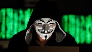 Person im schwarzen Kapuzenpullover sitzt mit Guy Fawkes Maske vor einem Laptop.