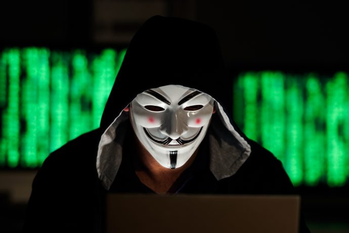 Person im schwarzen Kapuzenpullover sitzt mit Guy Fawkes Maske vor einem Laptop.