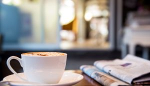 Kaffeetasse steht neben Zeitung auf einem Holztisch.