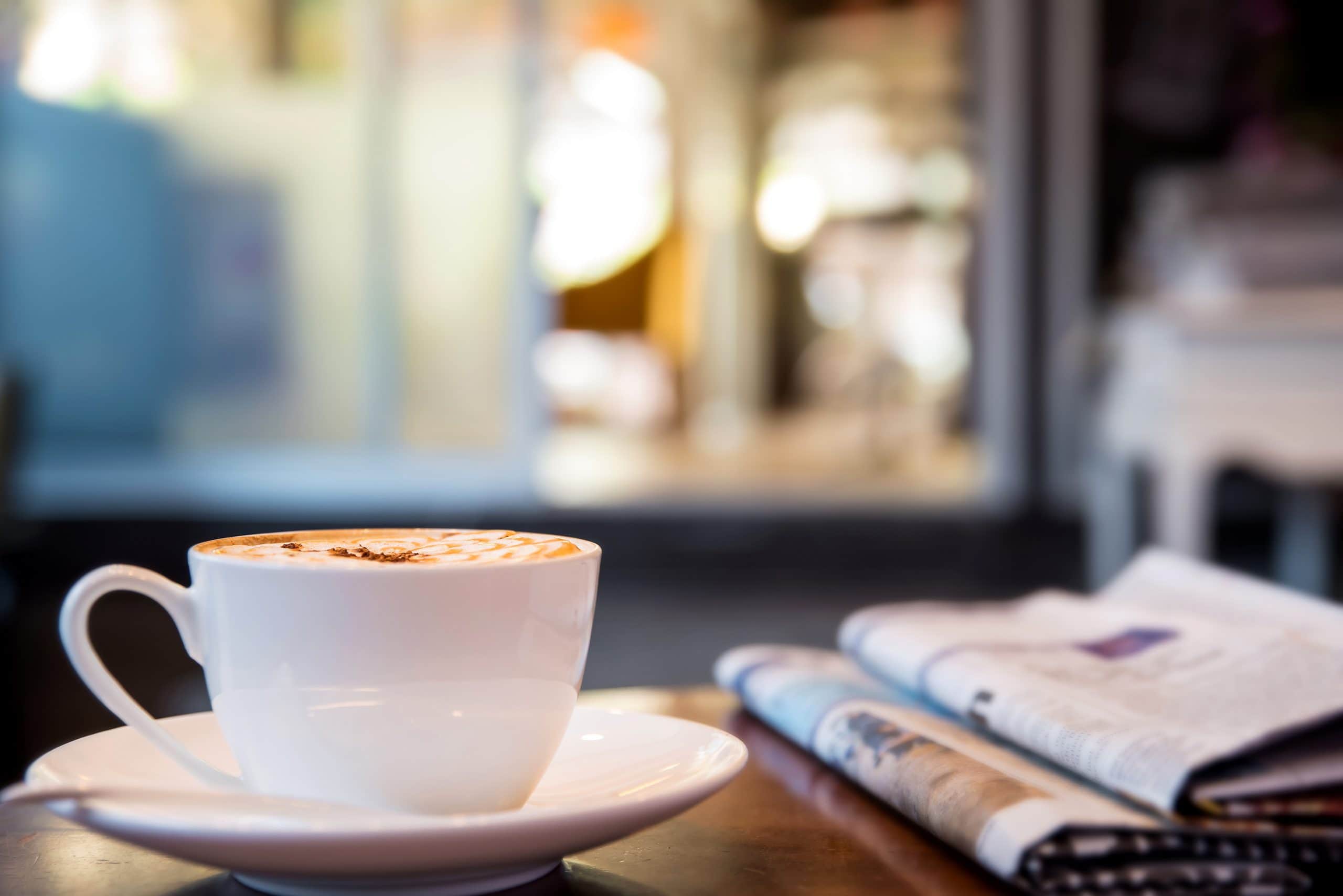 Kaffeetasse steht neben Zeitung auf einem Holztisch.