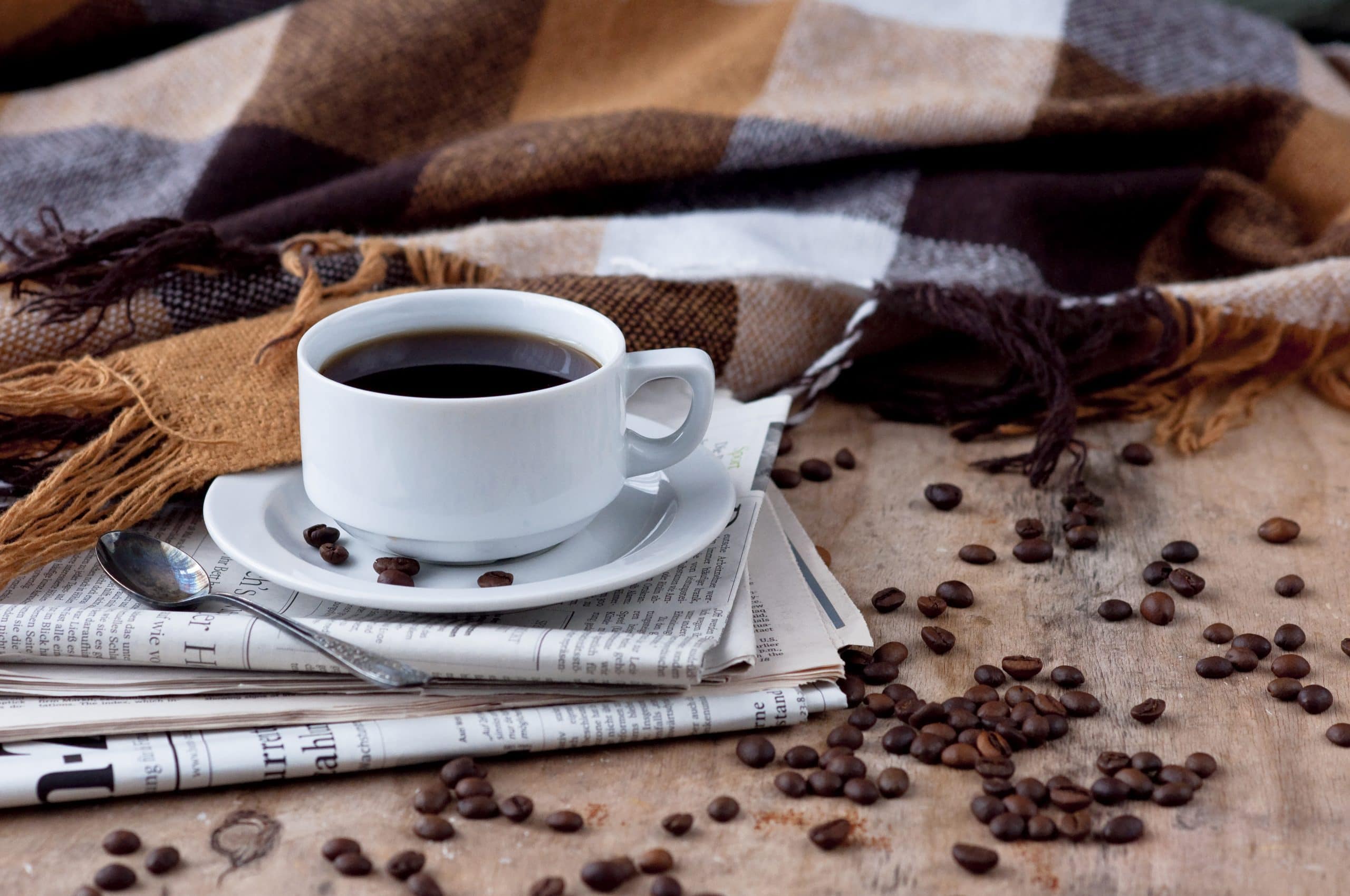 Eine weiße Kaffeetasse steht auf einem Stapel Zeitungen. Daneben liegen Kaffeebohnen und eine Wolldecke.