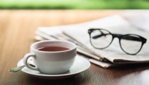 Eine Tasse Kaffee steht auf einem Holztisch neben einer Zeitung und eine Lesebrille.