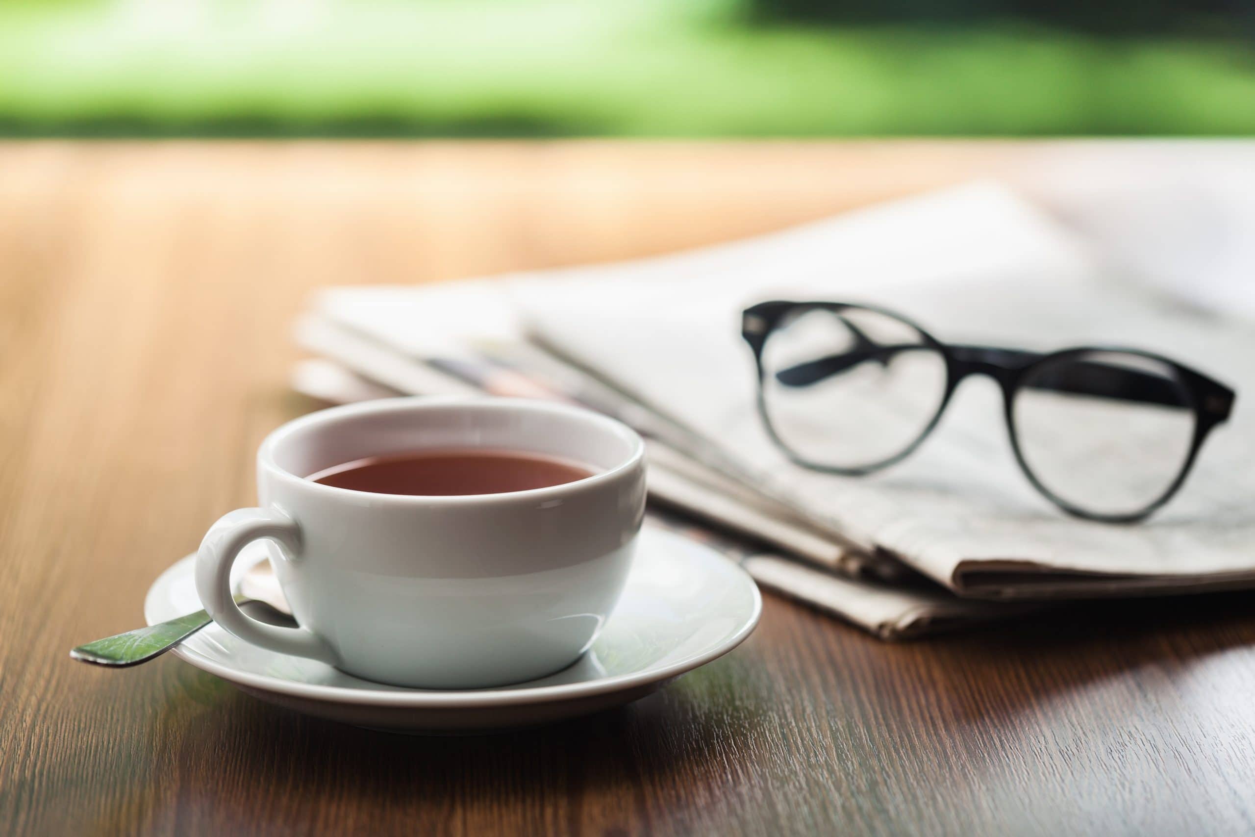 Eine Tasse Kaffee steht auf einem Holztisch neben einer Zeitung und eine Lesebrille.