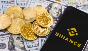 Krypto-Münzen liegen auf US-Dollar-Noten. Daneben liegt ein Smartphone mit Binance-Logo