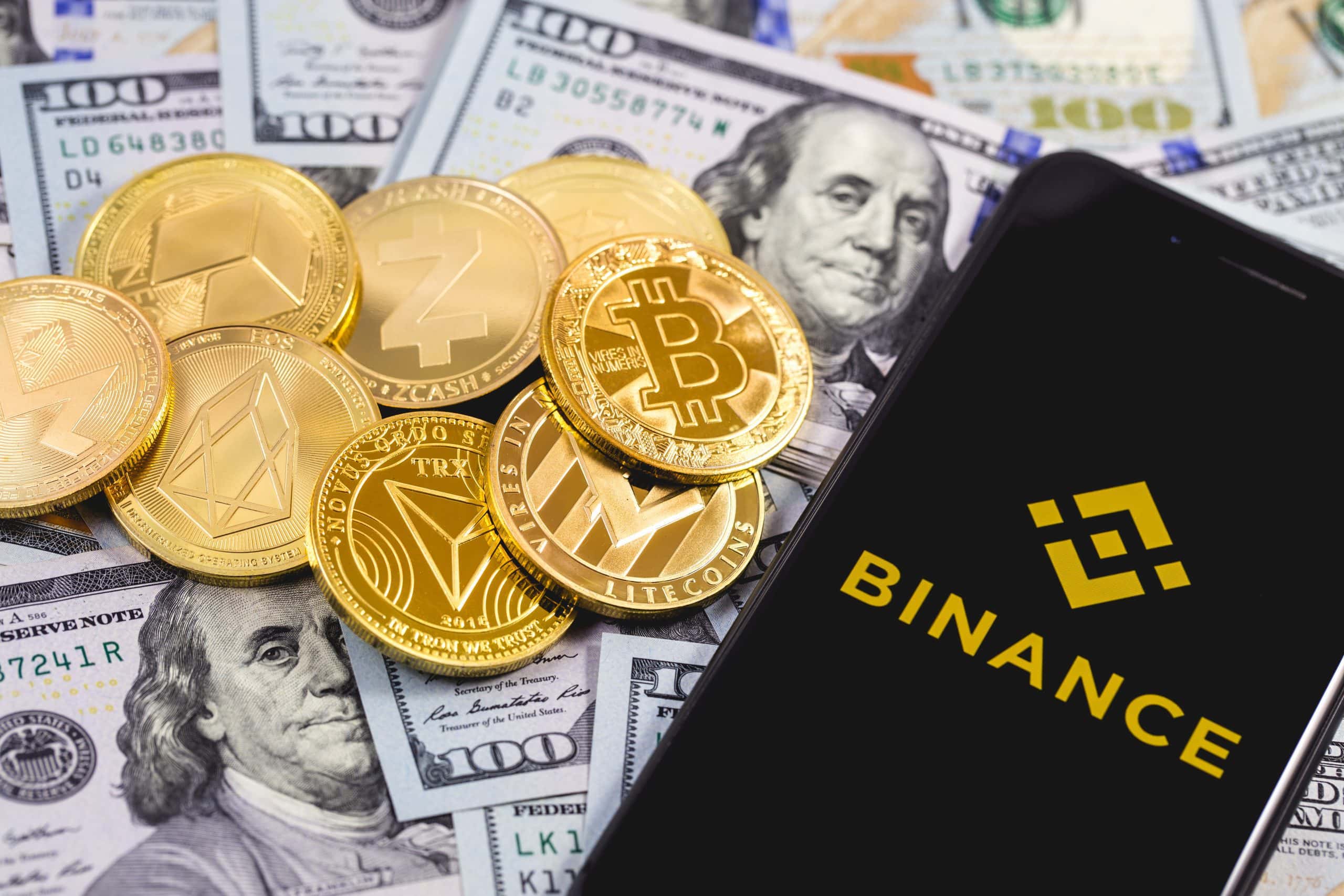 Krypto-Münzen liegen auf US-Dollar-Noten. Daneben liegt ein Smartphone mit Binance-Logo