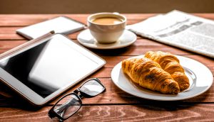 Kaffeetasse, Tablet, Brille, zwei Croissants, Handy und eine Zeitung auf einem Holztsich.