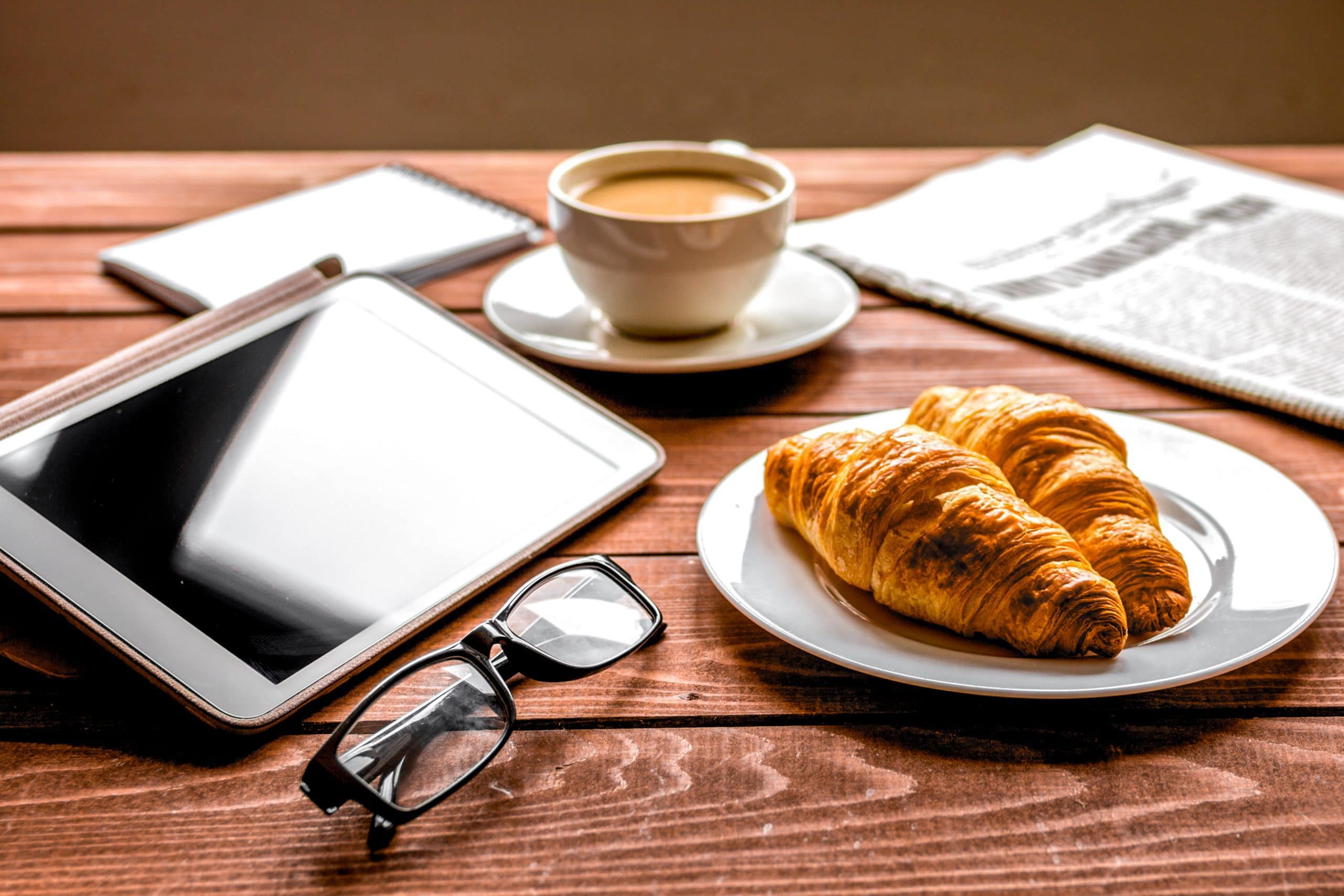 Kaffeetasse, Tablet, Brille, zwei Croissants, Handy und eine Zeitung auf einem Holztsich.