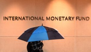 Logo des Internationalen Währungsfonds auf einer Betonwand. Davor ein Mensch mit blauem Regenschrim.