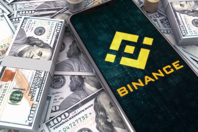 Geldbündel auf denen ein mobiles Endgerät liegt, dass auf der Seite von Binance ist