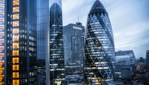 Moderne Wolkenkratzer im Londoner Finanzviertel, darunter das Gherkin und die Büros von Firmen wie Blossom Capital, werden in der Abenddämmerung mit leuchtenden Lichtern und schimmernden Spiegelbildern in den umliegenden Glasgebäuden gezeigt.