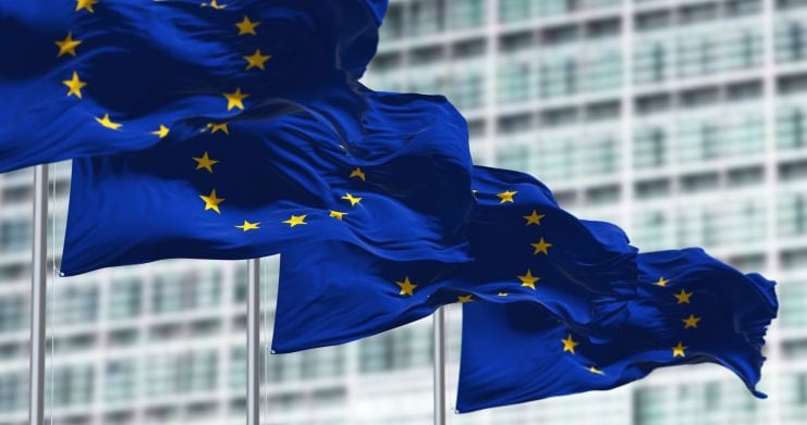 Drei Flaggen der Europäischen Union wehen anmutig vor einem modernen Glasgebäude und spiegeln die nahtlose Integration von Tradition und Innovation in der sich ständig weiterentwickelnden Welt digitaler Währungen wie Krypto wider.
