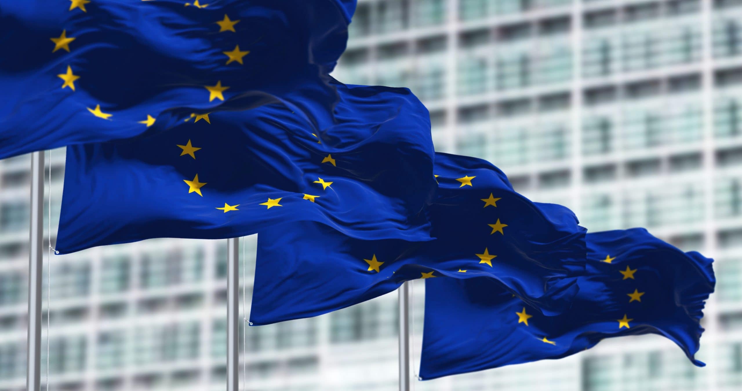 Drei Flaggen der Europäischen Union wehen anmutig vor einem modernen Glasgebäude und spiegeln die nahtlose Integration von Tradition und Innovation in der sich ständig weiterentwickelnden Welt digitaler Währungen wie Krypto wider.