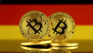 Zwei physische Bitcoin-Token stehen aufrecht vor einer deutschen Flagge und symbolisieren die Bekanntheit von Krypto. Weitere Münzen liegen flach darunter.