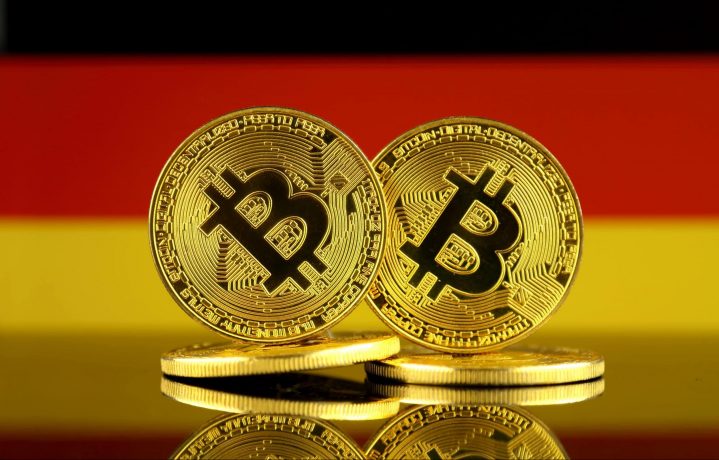 Zwei physische Bitcoin-Token stehen aufrecht vor einer deutschen Flagge und symbolisieren die Bekanntheit von Krypto. Weitere Münzen liegen flach darunter.