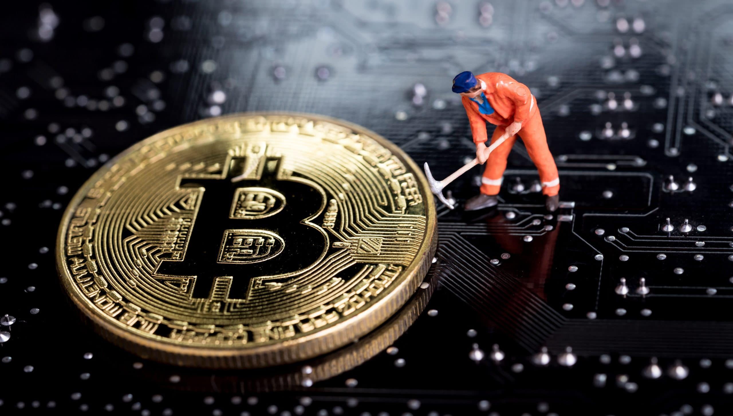 Eine Miniaturfigur in leuchtend orangefarbener Kleidung steht mit einer Spitzhacke in der Hand neben einer glänzenden Bitcoin-Münze auf einer Platine und fängt die Essenz des Kryptowährungs-Minings in Aktion perfekt ein.