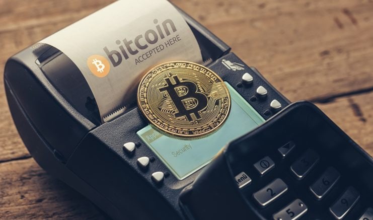 Eine Bitcoin-Münze liegt auf einem Kreditkartenlesegerät. Ihre Anwesenheit wird durch den Beleg hervorgehoben, auf dem steht: „Hier wird Bitcoin akzeptiert.“.