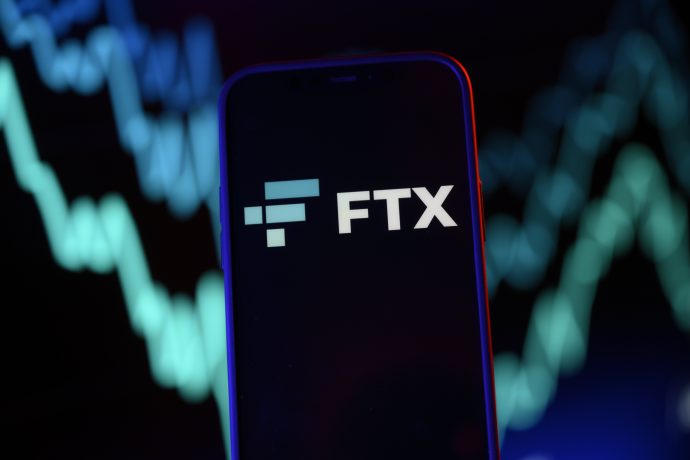 Auf einem Smartphone ist das FTX-Logo vor dem dynamischen Hintergrund blauer und grüner Finanzdiagrammlinien gut sichtbar.