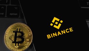 Eine schimmernde goldene Bitcoin-Münze liegt neben einem Smartphone-Bildschirm, auf dem stolz das Binance-Logo zu sehen ist, und das Ganze elegant auf einer dunklen Laptop-Tastatur positioniert.