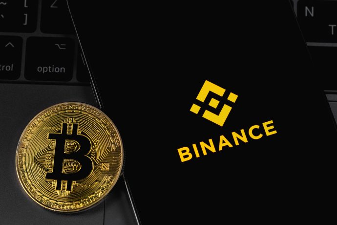 Eine schimmernde goldene Bitcoin-Münze liegt neben einem Smartphone-Bildschirm, auf dem stolz das Binance-Logo zu sehen ist, und das Ganze elegant auf einer dunklen Laptop-Tastatur positioniert.