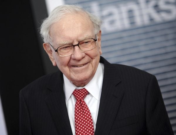 Warren Buffett in schwarzem Anzug, weißem Hemd und rot gepunkteter Krawatte steht lächelnd vor einem verschwommenen Hintergrund.