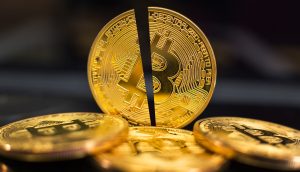 Eine in zwei Hälften geteilte goldene Bitcoin-Münze steht aufrecht zwischen anderen ganzen Bitcoin-Münzen auf einer dunklen Oberfläche und symbolisiert die Volatilität von Bitcoin.
