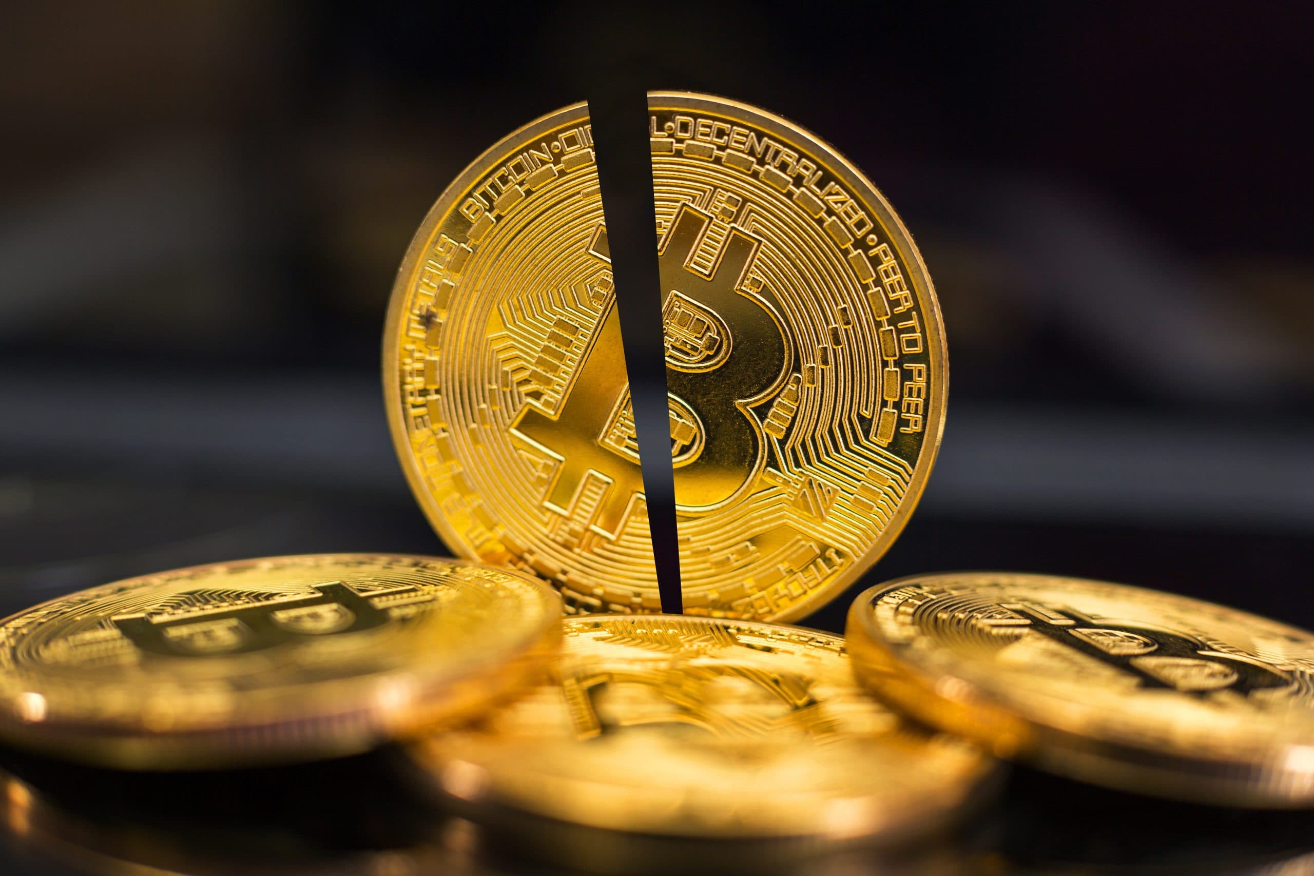 Eine in zwei Hälften geteilte goldene Bitcoin-Münze steht aufrecht zwischen anderen ganzen Bitcoin-Münzen auf einer dunklen Oberfläche und symbolisiert die Volatilität von Bitcoin.