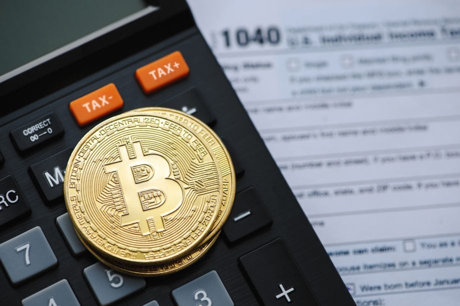 Eine goldene Bitcoin-Münze ruht auf einem Taschenrechner mit TAX-Tasten neben einem US-Einkommenssteuerformular 1040, um die Rolle von Bitcoin in der Steuersaison hervorzuheben.