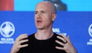 Ein kahlköpfiger Mann mit schwarzem Hemd und Headset-Mikrofon gestikuliert mit beiden Händen, während er auf der Bühne vor einem blauen Hintergrund mit Coinbase-Logos und weißem Text spricht.